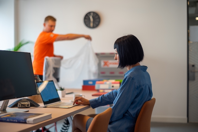 A woman working on a laptop while a worker unpacks items in the background.
