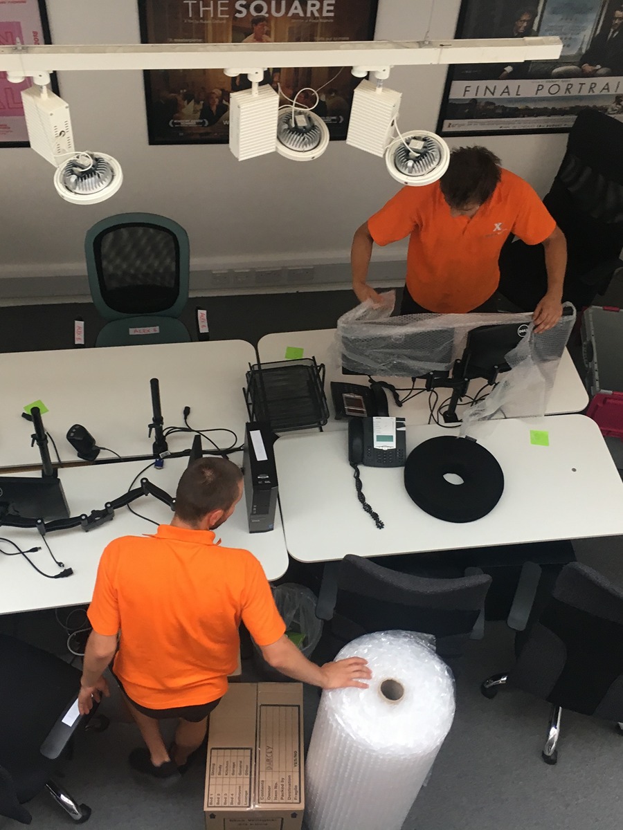 A top-down view of two workers packing office equipment with boxes and bubble wrap.